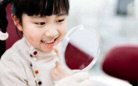 Asian girl looking at her teeth in the mirror and sitting at dentist’s chair.