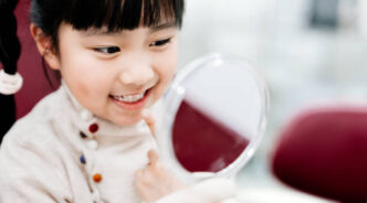 Asian girl looking at her teeth in the mirror and sitting at dentist’s chair.